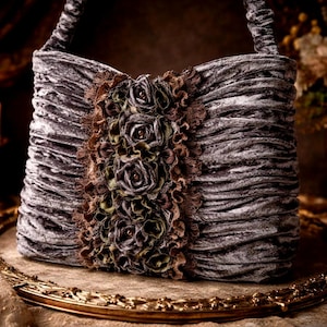 Könnte beinhalten: Graue Samt-Handtasche mit gerüschtem Design und floraler Verzierung. Die Tasche hat einen Griff und wird auf einem goldfarbenen Tablett präsentiert, mit Kerzen und Blumen im Hintergrund.