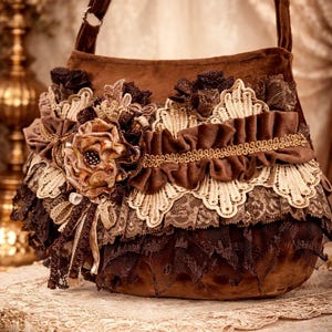 Può includere: Una borsa in camoscio marrone con tracolla, ornata da strati di pizzo, balze e abbellimenti floreali. La borsa presenta un grande fiore in tessuto e finiture intricate, creando un'estetica vintage e romantica. La palette di colori comprende tonalità di marrone e crema.
