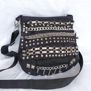Könnte beinhalten: Schwarze Umhängetasche mit silbernen Metallketten, Nieten und Stacheln. Die Tasche hat einen verstellbaren schwarzen Riemen und einen silbernen Metallverschluss. Die Tasche besteht aus schwarzem Stoff mit einem Used-Look.