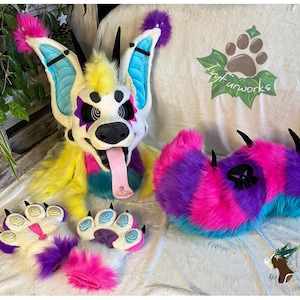 Può includere: Un set di fursuit colorato composto da testa, zampe e coda. La testa è bianca con dettagli gialli, blu e viola, e una lunga lingua rosa. Le zampe e la coda presentano una vivace pelliccia rosa, viola e blu.