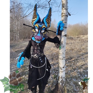 May include: A furry costume featuring a detailed, stylized wolf or fox head with large ears, blue and brown accents, and a purple muzzle. The costume includes matching blue paw gloves and a black outfit with a chain belt. The background is a natural outdoor setting.