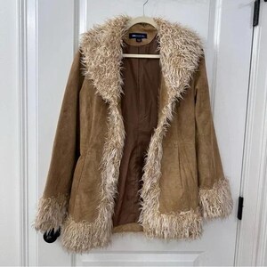 May include: A tan suede jacket with a faux fur collar, cuffs, and trim. The jacket is hanging on a wooden hanger against a white door. The jacket has a button closure and a brown lining.