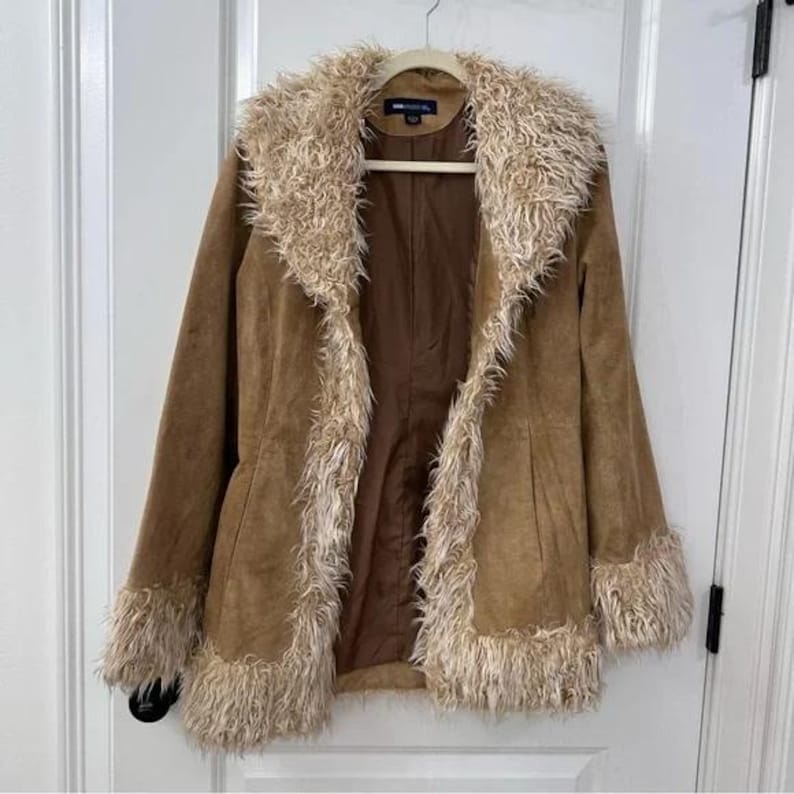 May include: A tan suede jacket with a faux fur collar, cuffs, and trim. The jacket is hanging on a wooden hanger against a white door. The jacket has a button closure and a brown lining.
