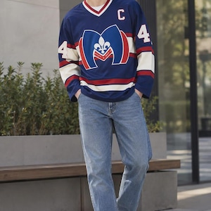 May include: A navy blue long-sleeve jersey with a white V-neck collar featuring red stripes. The front displays a large blue letter M with a white fleur-de-lis, the letter C, and the number 4 on the chest and sleeves. Paired with blue denim jeans.
