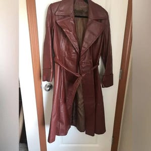 May include: A long, reddish-brown leather trench coat with a matching belt. The coat has a wide collar, long sleeves, and a tailored fit. It is hanging on a black hanger on a white door.