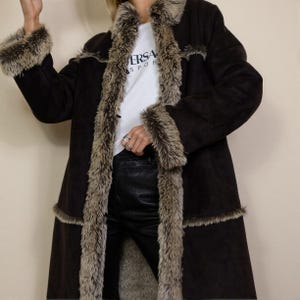 May include: A long, dark brown coat with a soft, light brown faux fur trim along the collar, front opening, cuffs, and horizontal seams. The coat is worn over a white t-shirt with the text VERSACE SPORT and black trousers.