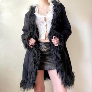 May include: Black faux fur trim coat. The coat has a longline silhouette with a faux fur collar and cuffs. The coat is worn over a white top and a black leather skirt. A silver belt with a decorative buckle is also visible.