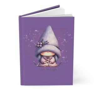 Puede incluir: Un diario morado con una ilustración caprichosa de un gnomo con un sombrero y abrigo lavanda. El gnomo está decorado con flores y el texto "I am enough" está en la parte inferior. El diario está abierto, revelando páginas blancas.