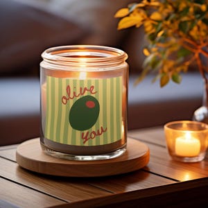 Può includere: Una candela accesa in un barattolo di vetro con un'etichetta che recita "alive You" con una grafica di oliva. La candela è appoggiata su un sottobicchiere di legno. Anche una piccola candela in un portacandele di vetro è accesa.