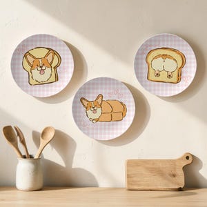 May include: Three decorative plates with corgi dog illustrations. Each plate features a different corgi design, including a corgi in a slice of bread, a corgi's rear end in bread, and a corgi lying down. The plates have a pink and white checkered background.