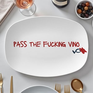 May include: White oval serving platter with the text "PASS THE FUCKING VINO" in red. A small graphic of a wine glass and spilled wine is on the right. The platter is surrounded by wine glasses, a bottle of wine, and small bowls.
