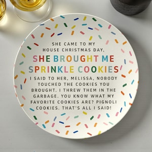 May include: A white ceramic plate with colorful sprinkles and text. The text reads "SHE BROUGHT ME SPRINKLE COOKIES!" and continues with a humorous message about cookies. The plate is round and has a slightly raised edge.