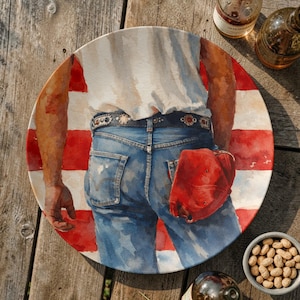 May include: A round decorative plate featuring a watercolor painting of a person in blue jeans and a red baseball cap. The plate has a red and white striped background, reminiscent of the American flag.
