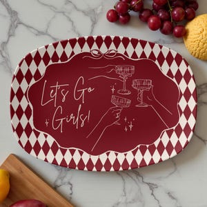 May include: An oval serving platter with a burgundy center and a white diamond pattern border. The center features the text "Let's Go Girls!" with illustrations of hands toasting with champagne glasses. A fun and festive design.