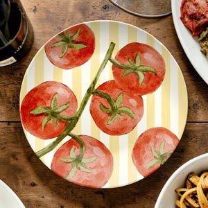 May include: Round ceramic plate with a yellow and white striped background. The plate features a watercolor illustration of red tomatoes on a green vine. The tomatoes have green stems and leaves. The plate is on a wooden surface.