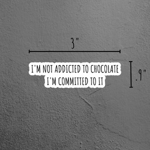 May include: A white sticker with black text that reads "I'm not addicted to chocolate I'm committed to it". The sticker is on a gray textured surface. The sticker is 3 inches wide and 9 inches tall.