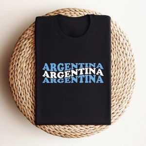 Puede incluir: Una camiseta negra con la palabra "ARGENTINA" repetida tres veces en una fuente retro apilada. El texto es blanco con acentos azules.