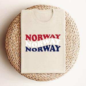 May include: A beige t-shirt with the word "NORWAY" printed three times in red, white, and blue. The word is stacked on top of itself, with the red letters on top, the white letters in the middle, and the blue letters on the bottom.
