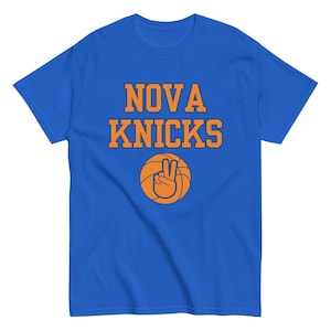 May include: Royal blue t-shirt with the words "NOVA KNICKS" in orange block letters. Below the text is an orange basketball with a hand making a peace sign. The shirt is a crew neck style.