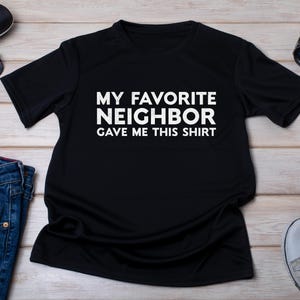 Op de afbeelding: Zwart T-shirt met witte tekst die luidt: "Mijn favoriete buurman heeft me dit shirt gegeven."