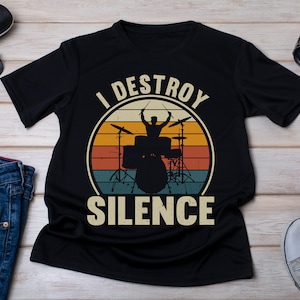 Pode incluir: Camiseta preta com um gráfico de estilo vintage de um baterista tocando uma bateria. O gráfico é rodeado por um círculo com o texto "I DESTROY SILENCE".