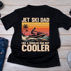 May include: Black t-shirt with the text "JET SKI DAD LIKE A REGULAR DAD BUT COOLER" and a graphic of a person riding a jet ski at sunset. Sunglasses, a watch, and sneakers are also visible.