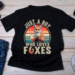 Peut inclure: T-shirt noir avec un motif de renard dans un cercle, avec le texte "JUST A BOY WHO LOVES FOXES". Le renard est brun et blanc, et le motif a des rayures orange, rouge et turquoise.