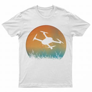 May include: White t-shirt with a graphic of a drone flying over a field of grass. The graphic is a circle with a sunset gradient of orange, yellow, and blue.