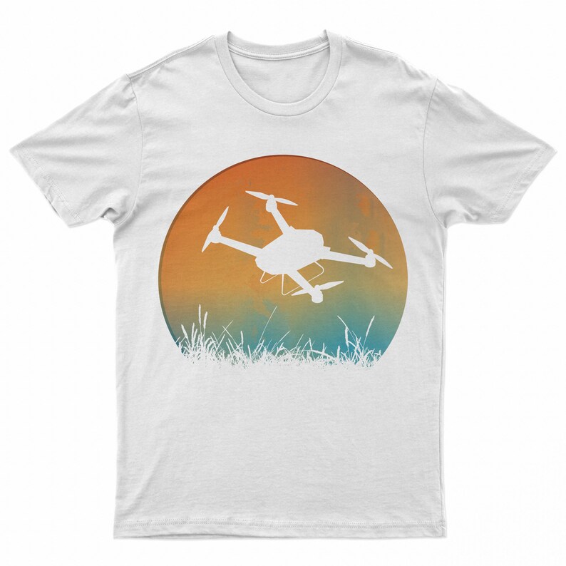 May include: White t-shirt with a graphic of a drone flying over a field of grass. The graphic is a circle with a sunset gradient of orange, yellow, and blue.