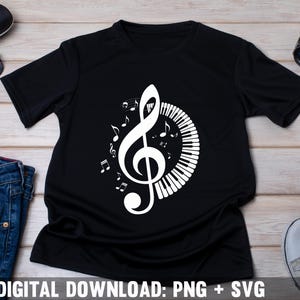 Op de afbeelding: Zwart T-shirt met een wit ontwerp van een vioolsleutel en pianotoetsen. Het ontwerp bevat muzieknoten rond de vioolsleutel.