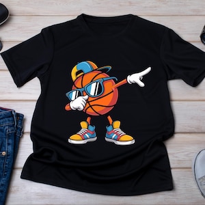 May include: Black t-shirt with a cartoon basketball character dabbing, wearing sunglasses and a cap. The character is orange with white gloves and colourful trainers. Sunglasses, a watch, and jeans are also visible.