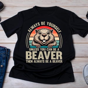 May include: A black short-sleeved t-shirt featuring a graphic of a beaver centred within a circular sunset design. The text reads: ALWAYS BE YOURSELF UNLESS YOU CAN BE A BEAVER THEN ALWAYS BE A BEAVER. The design uses orange, red, and teal colours.