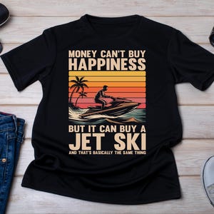 May include: Black t-shirt featuring a jet ski graphic with a sunset backdrop and the text: "MONEY CAN'T BUY HAPPINESS BUT IT CAN BUY A JET SKI AND THAT'S BASICALLY THE SAME THING."