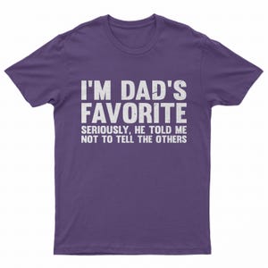 May include: A purple t-shirt with the text "I'M DAD'S FAVORITE" in large white letters. Below, smaller text reads "SERIOUSLY, HE TOLD ME NOT TO TELL THE OTHERS". The shirt is a casual wear item.