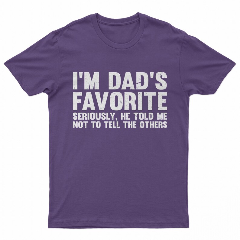 May include: A purple t-shirt with the text "I'M DAD'S FAVORITE" in large white letters. Below, smaller text reads "SERIOUSLY, HE TOLD ME NOT TO TELL THE OTHERS". The shirt is a casual wear item.