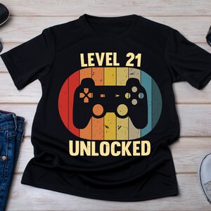 May include: Black t-shirt with a retro-style graphic of a video game controller and the text "LEVEL 21 UNLOCKED".