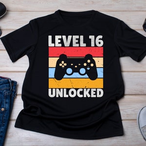 May include: Black t-shirt with a retro-style graphic of a video game controller and the text "Level 16 Unlocked".
