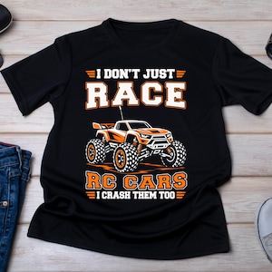 Könnte beinhalten: Schwarzes T-Shirt mit dem Text "I DON'T JUST RACE RC CARS I CRASH THEM TOO" und einer orange-weißen Grafik eines RC-Cars. Das Shirt wird mit Sonnenbrille, Uhr, Etui, Jeans und Turnschuhen auf einer Holzoberfläche präsentiert.