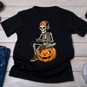 Op de afbeelding: Een zwart T-shirt met korte mouwen en een grafische print van een skelet met een oranje honkbalpet en een koptelefoon, zittend op een uitgesneden Halloween-pompoen terwijl het een videogame speelt met een controller.