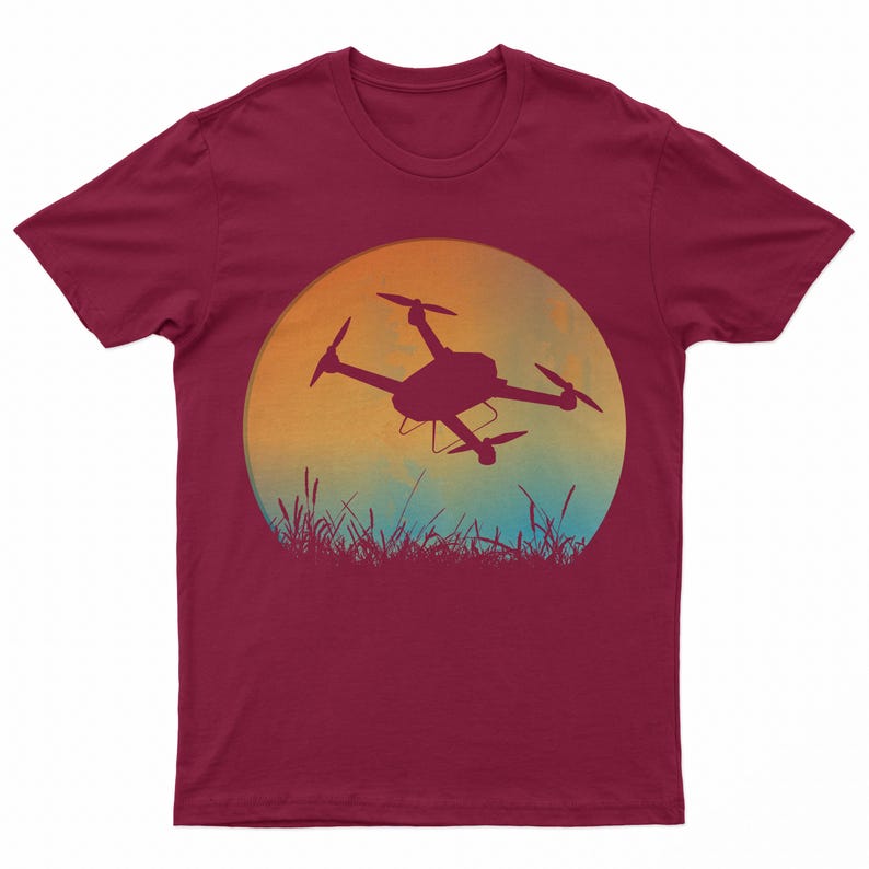 May include: A maroon t-shirt with a graphic of a drone flying in front of a sunset. The drone is a silhouette against a yellow, orange, and blue sky. The graphic is surrounded by a circle of grass.