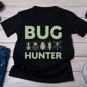 May include: Black t-shirt with the text "BUG HUNTER" in light green, featuring insect illustrations. The shirt is displayed with sunglasses, a watch, and jeans.