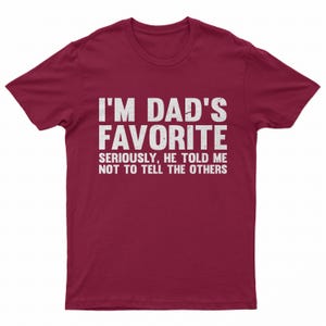 May include: A maroon t-shirt with white text that reads "I'M DAD'S FAVORITE" in a bold font. Below, in a smaller font, it says "SERIOUSLY, HE TOLD ME NOT TO TELL THE OTHERS."
