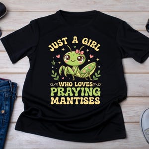 Peut inclure: Un t-shirt noir à manches courtes avec un motif de mante religieuse verte portant un nœud rose. Le texte "JUST A GIRL WHO LOVES PRAYING MANTISES" est écrit en jaune et vert, entouré de petits cœurs roses et de motifs de feuilles vertes.