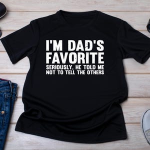 May include: Black t-shirt with the text "I'M DAD'S FAVORITE SERIOUSLY, HE TOLD ME NOT TO TELL THE OTHERS" in white. The shirt is displayed with sunglasses, a watch, a pair of jeans, and sneakers.