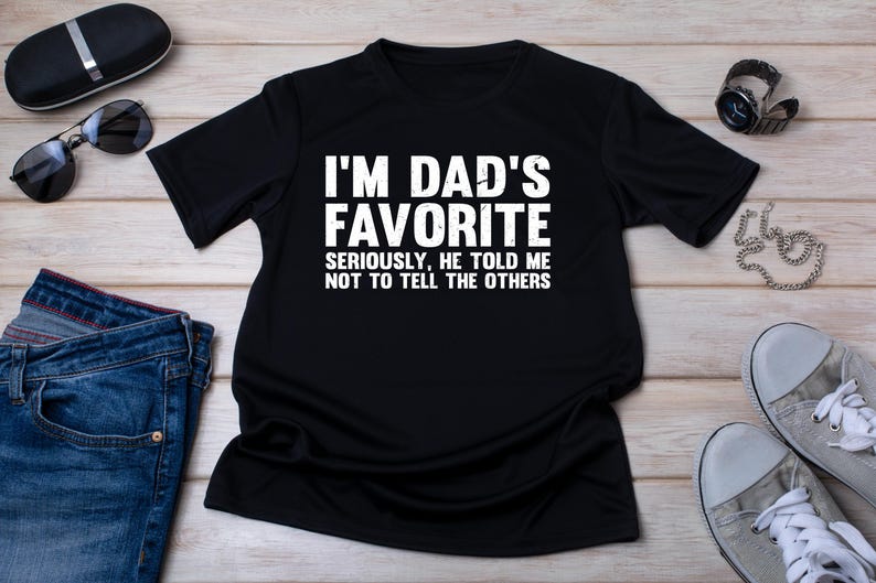 May include: Black t-shirt with the text "I'M DAD'S FAVORITE SERIOUSLY, HE TOLD ME NOT TO TELL THE OTHERS" in white. The shirt is displayed with sunglasses, a watch, a pair of jeans, and sneakers.