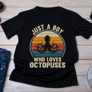 Op de afbeelding: Zwart t-shirt met een retro octopus ontwerp en de tekst "JUST A BOY WHO LOVES OCTOPUSES". Het shirt wordt getoond met een zonnebril, jeans en sneakers, op een houten oppervlak.