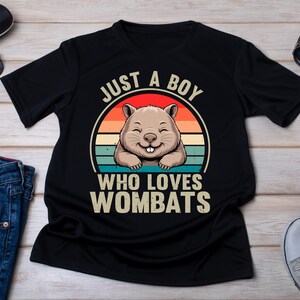 May include: Black t-shirt with a graphic of a smiling wombat inside a circular design with a sunset background. Text reads "JUST A BOY WHO LOVES WOMBATS". The shirt is displayed with sunglasses, jeans, and sneakers.