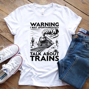 Puede incluir: Camiseta blanca con un gráfico en blanco y negro de un tren sobre vías, con el texto "WARNING I MAY SPONTANEOUSLY TALK ABOUT TRAINS." La camiseta se muestra con vaqueros azules y zapatillas blancas sobre una superficie de madera.