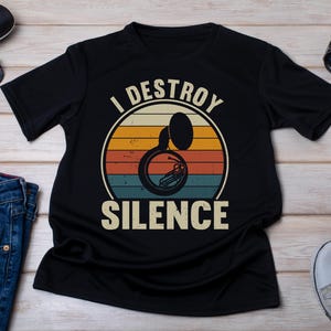 Peut inclure: Un t-shirt noir à manches courtes arborant un graphisme de soubassophone centré dans un design de coucher de soleil circulaire avec des rayures horizontales jaunes, orange et bleues. Le texte "I DESTROY SILENCE" est imprimé en lettres grasses.