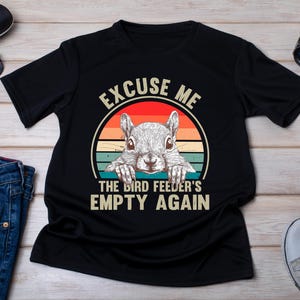 May include: Black t-shirt with a squirrel graphic inside a circle with a sunset design. The text reads "EXCUSE ME THE BIRD FEEDER'S EMPTY AGAIN". Other items include sunglasses, a watch, and jeans.